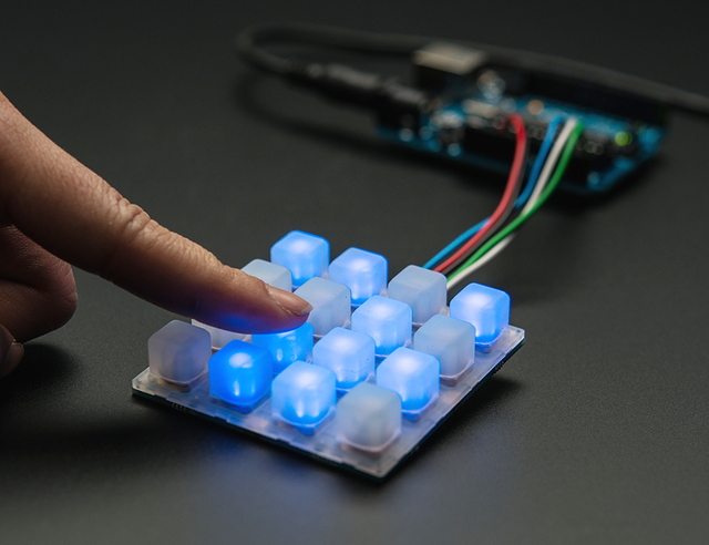 The silicone 4×4 keypad lit blue on a Trellis board, a finger pressing one key.