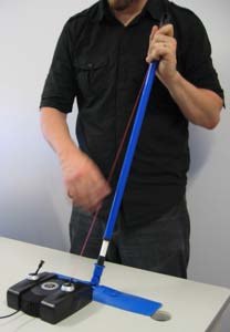 A player holds a long blue pole angled up from a table. Its foot is taped down beside a Gametrak, and a red tether runs from the Gametrak up along the pole to the player's upper hand, while the other hand plucks it.